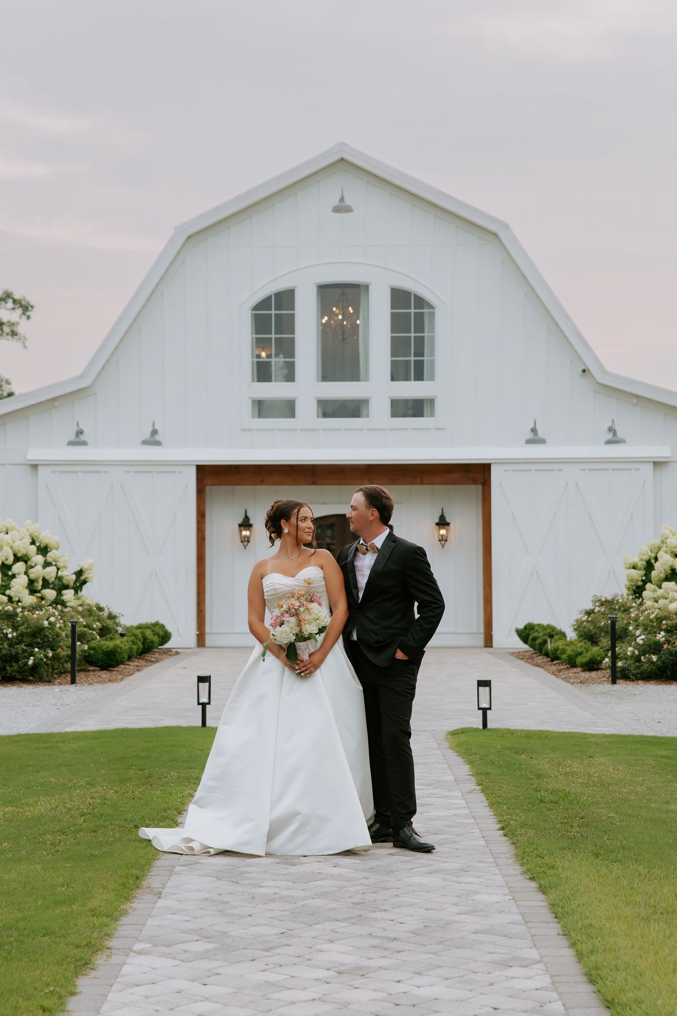 Bride and groom pose before a white barn wedding venue surrounded by lush greenery and hydrangeas at dusk at Heyward Manor.