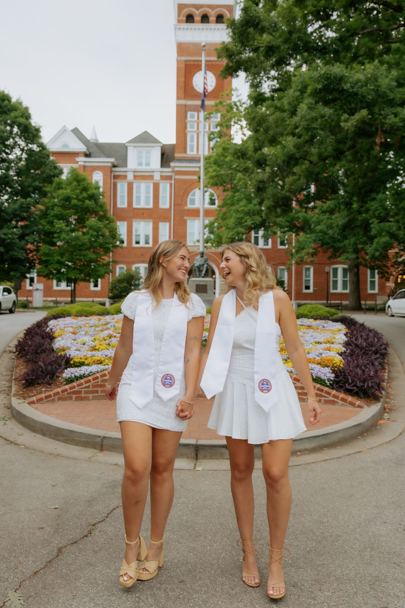 Two female graduates in white dresses with stoles laughing together in front of Clemson University building.