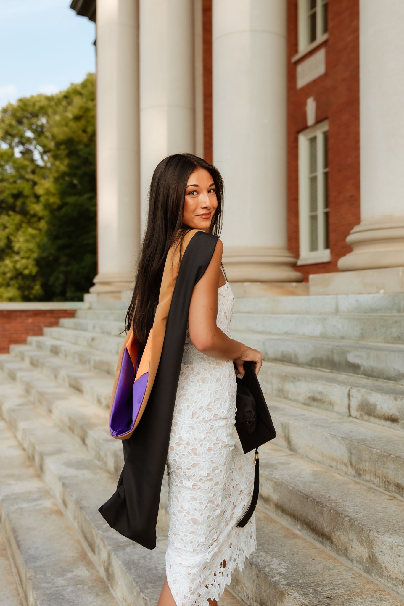 Graduate in white lace dress holding cap and gown on steps of brick Clemson University building.