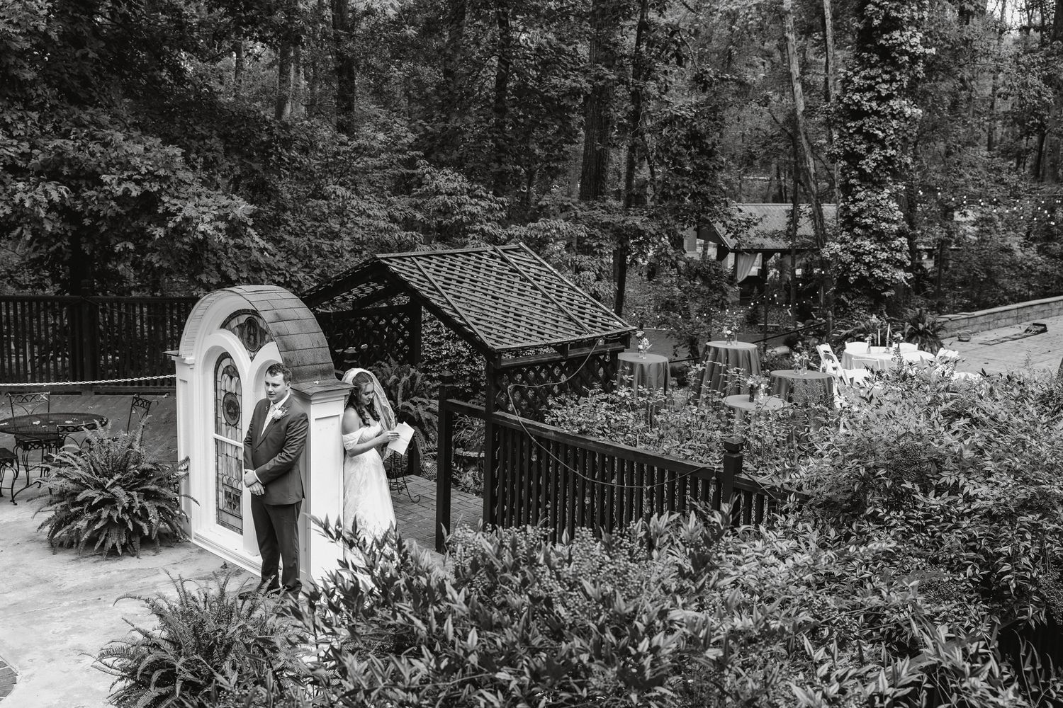 Black and white photo of bride and groom standing near a garden arch in a lush wooded setting at Lionsgate Manor.