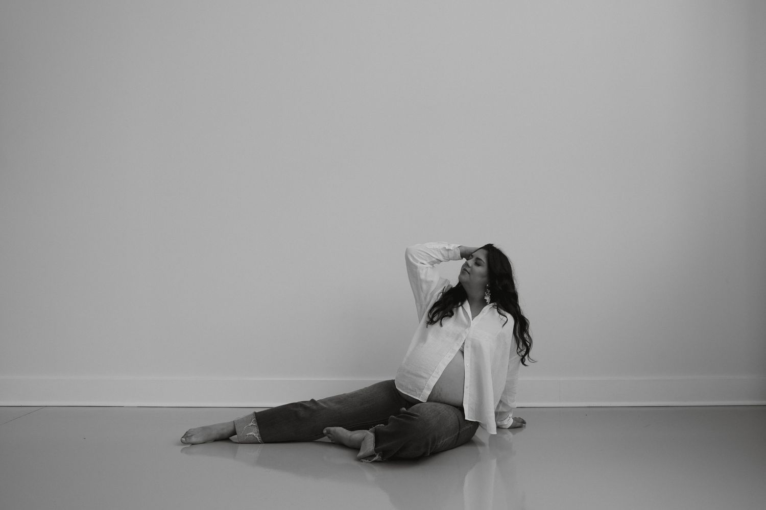 Black and white photo of woman sitting on floor posing in casual white shirt and jeans at WhiteWall Studios.