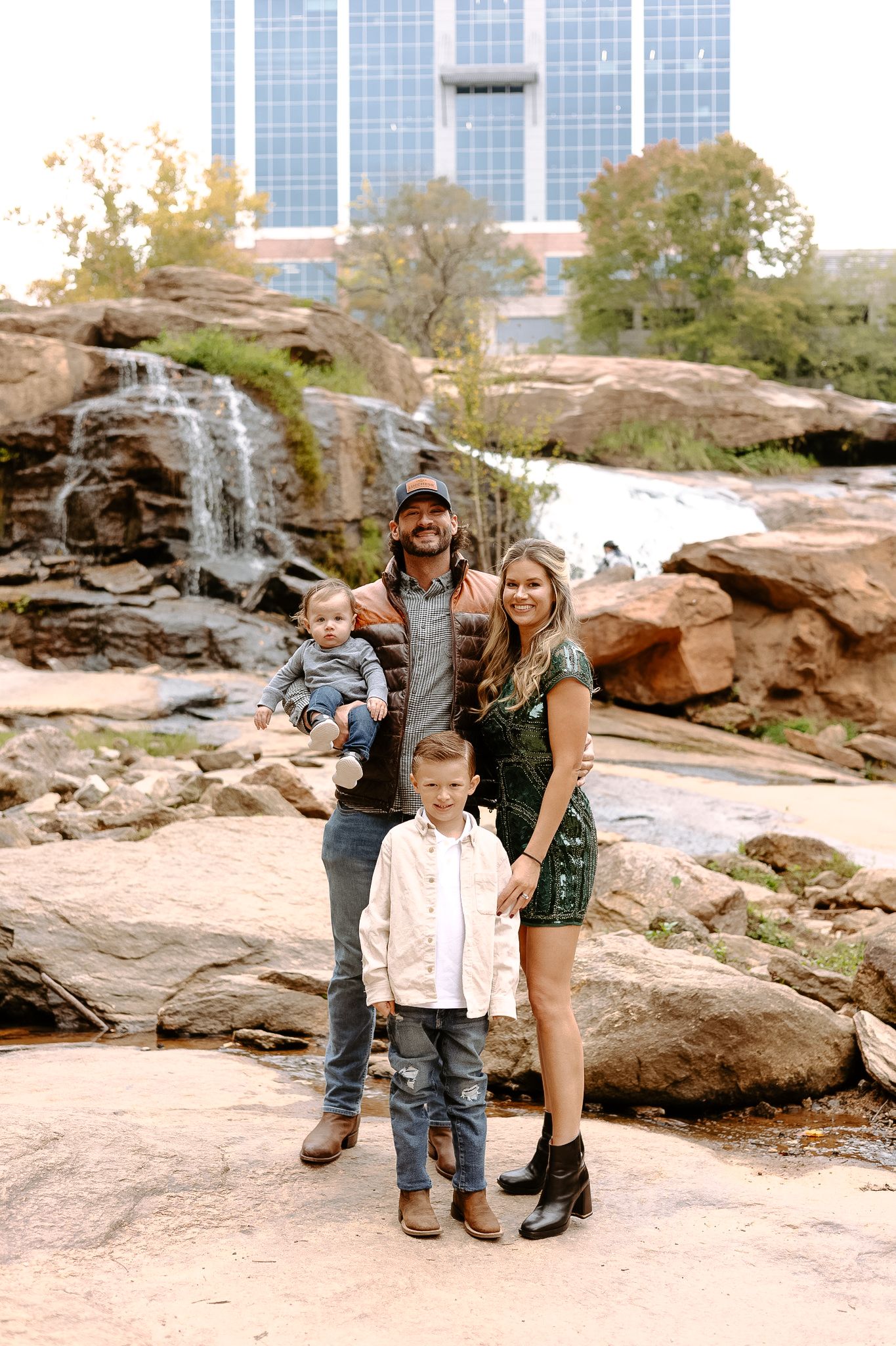 Family of four poses on rocky terrain near a waterfall with a city skyline in the background at Falls Park in Greenville.