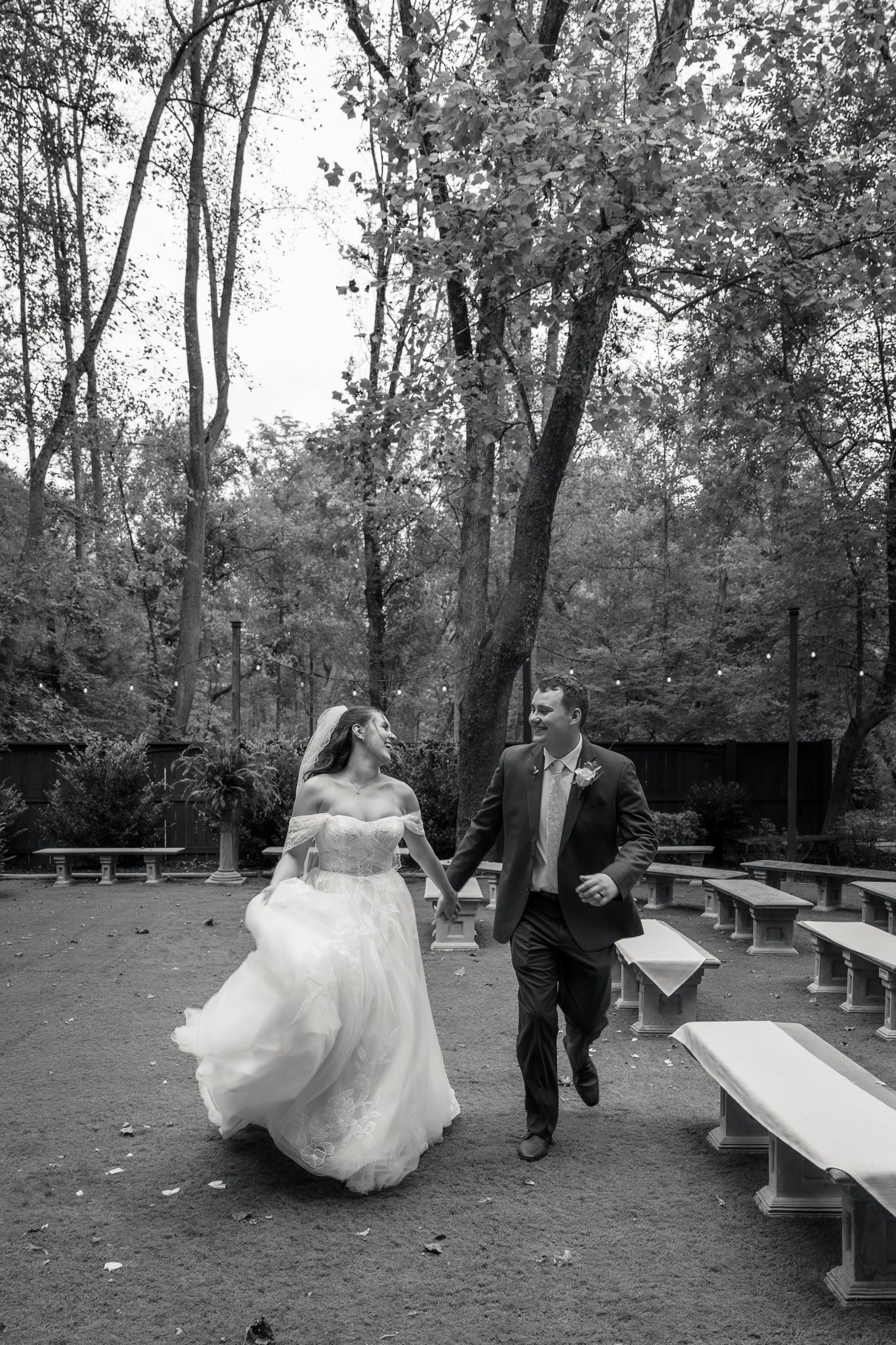 Black and white photo of bride and groom running hand-in-hand through an outdoor wooded ceremony space at Lionsgate Manor.