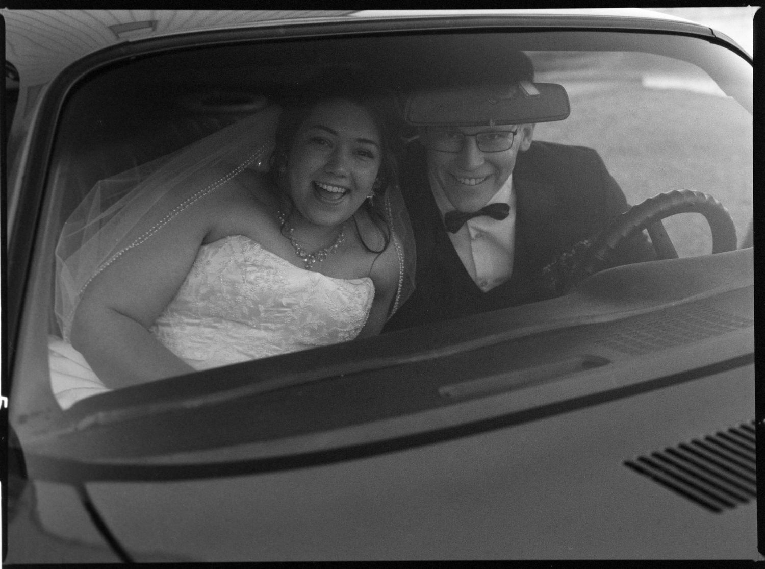 ©AnnaVonRiesenPhotography Wedding Photography | Film B&W image of the bride & groom laughing at the camera through the windshield of a car.