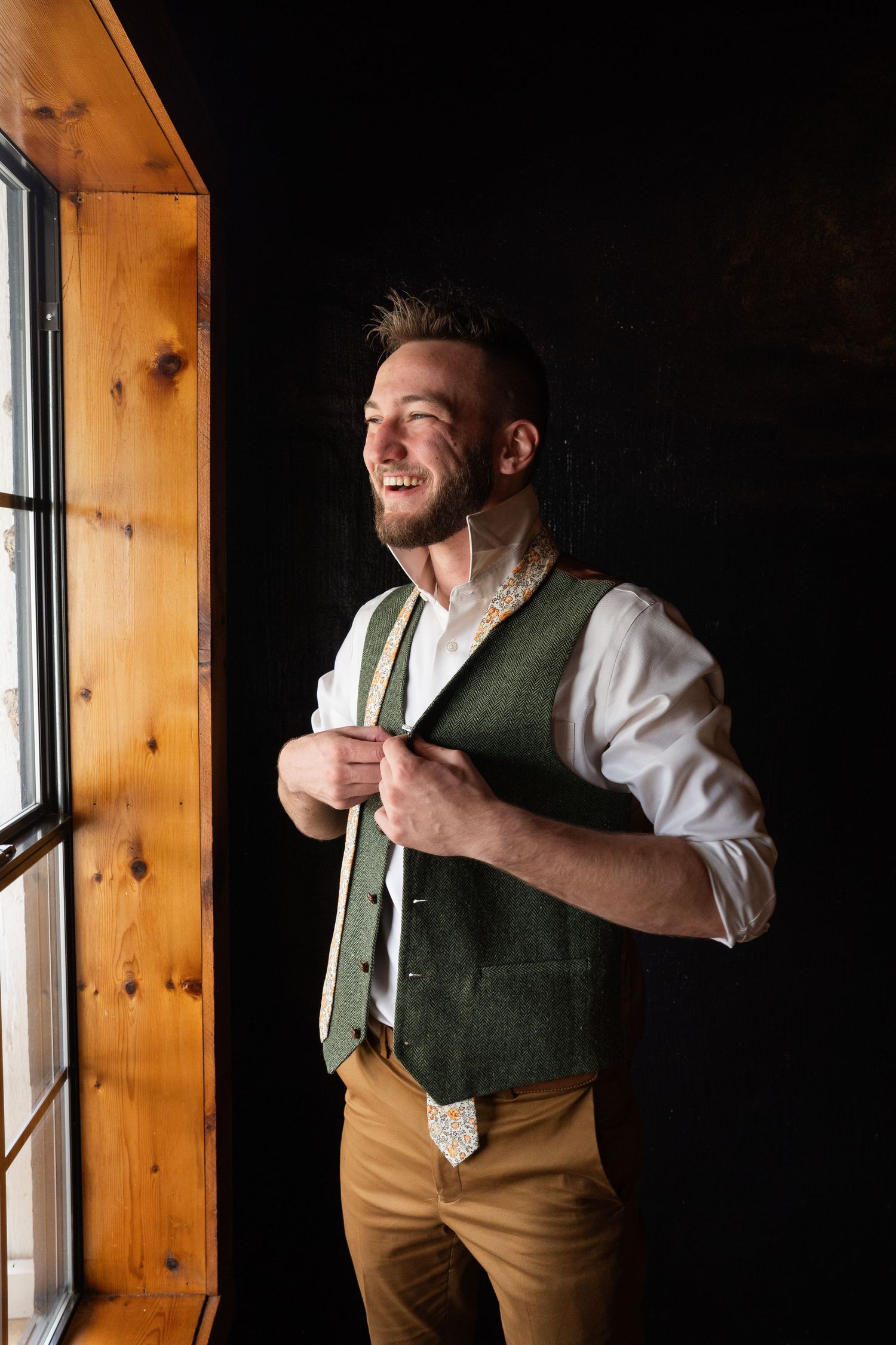 ©AnnaVonRiesenPhotography Wedding Photography | Color image of the groom smiling, standing by a window, tying his tie.