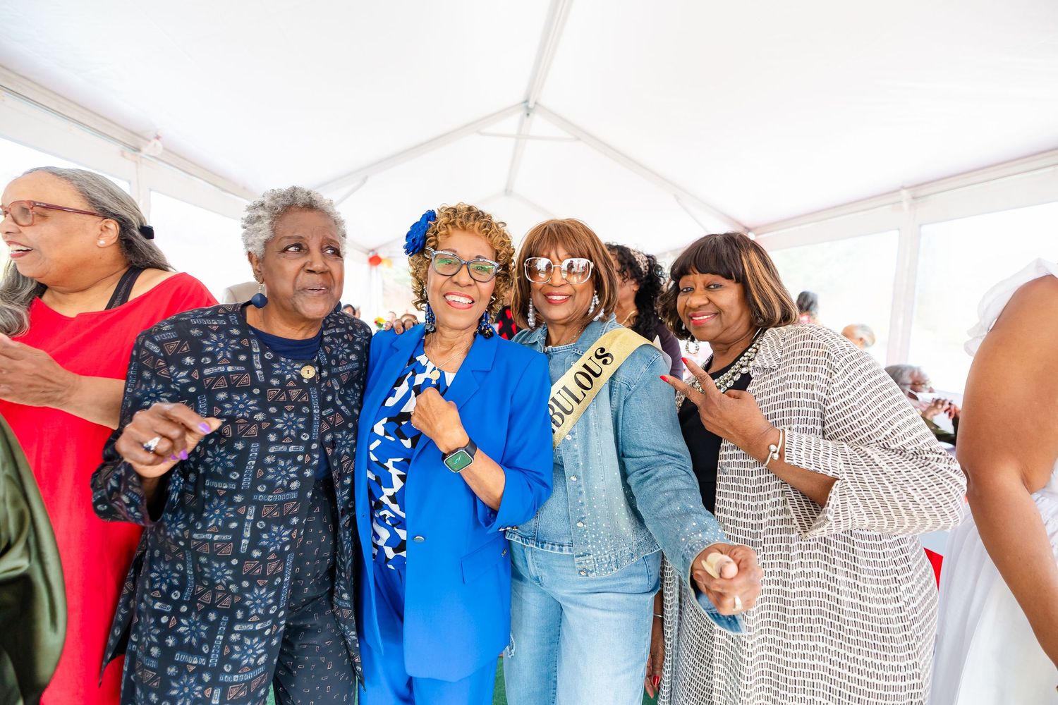 Group of five women smiling and posing together under a tent at an outdoor event.