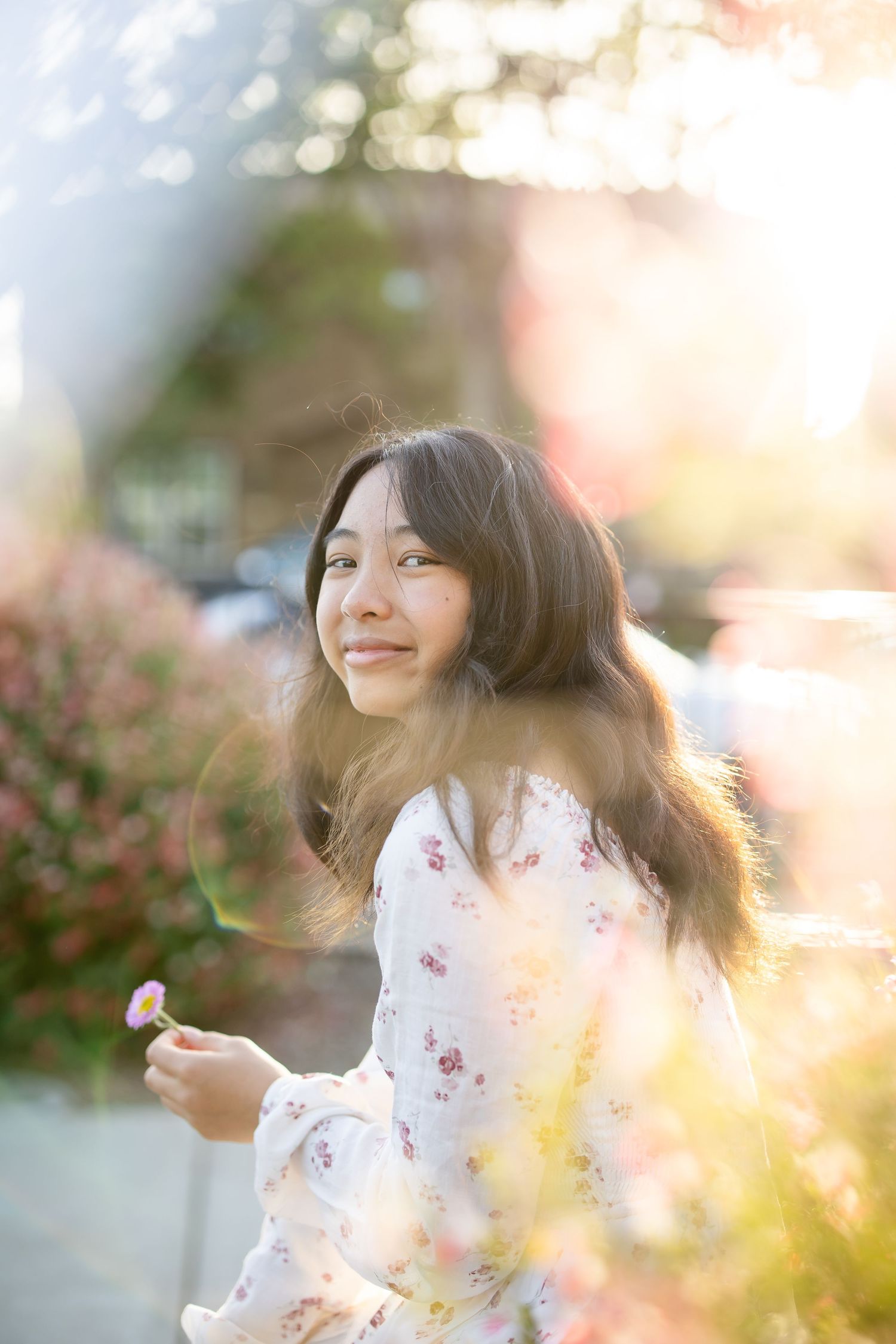 Young Asian woman in floral dress holding a flower in warm golden sunlight.