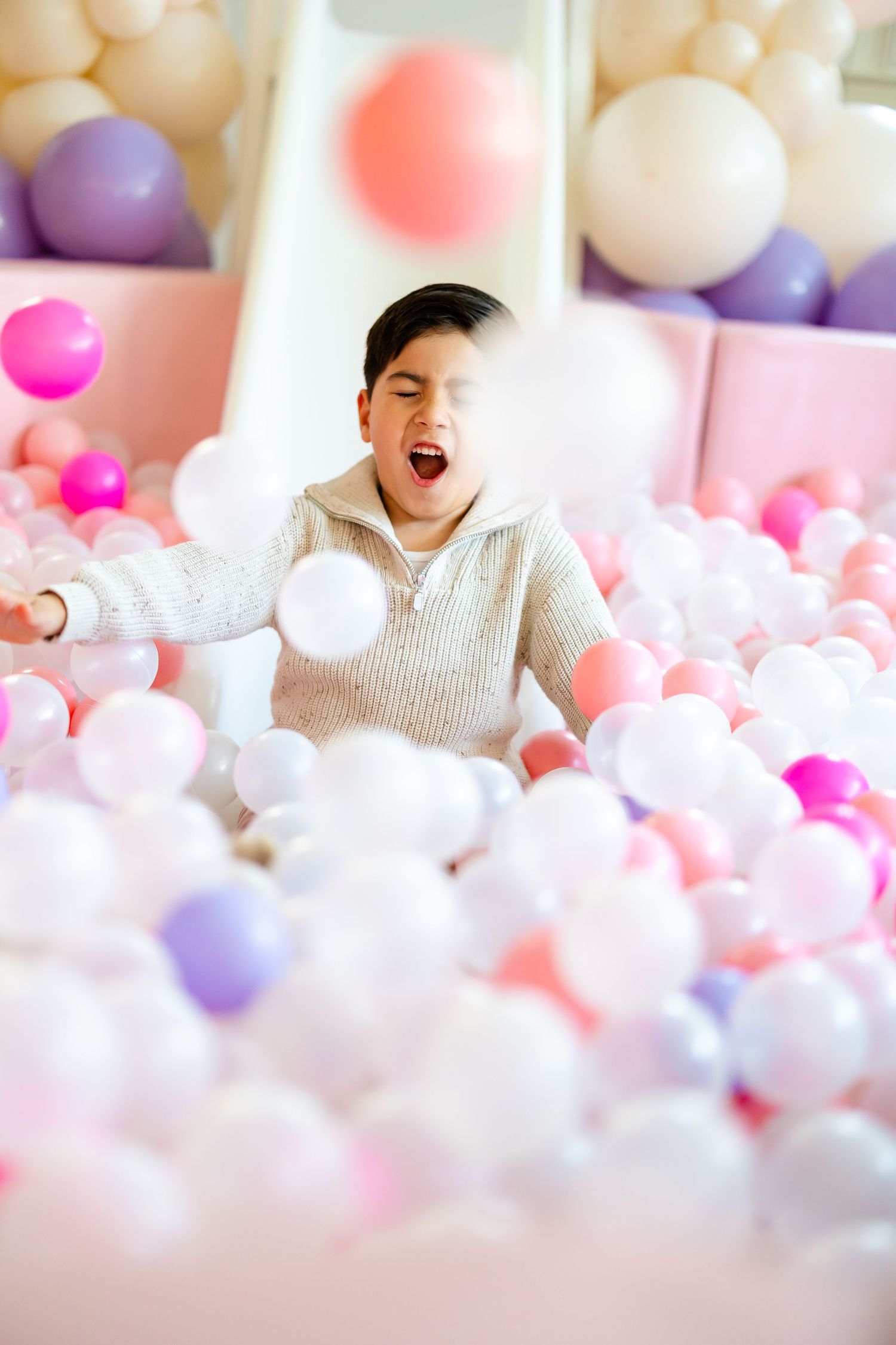 Excited child playing in a colorful pink and white ball pit with a slide.