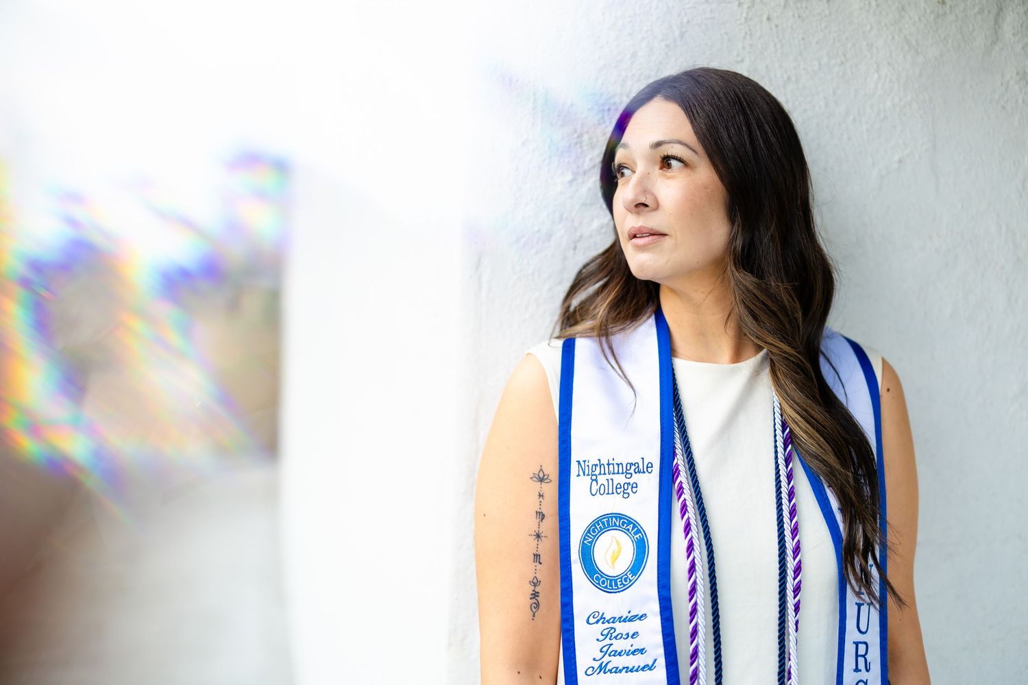 Woman in white dress wearing Nightingale College graduation sash leaning against a wall.