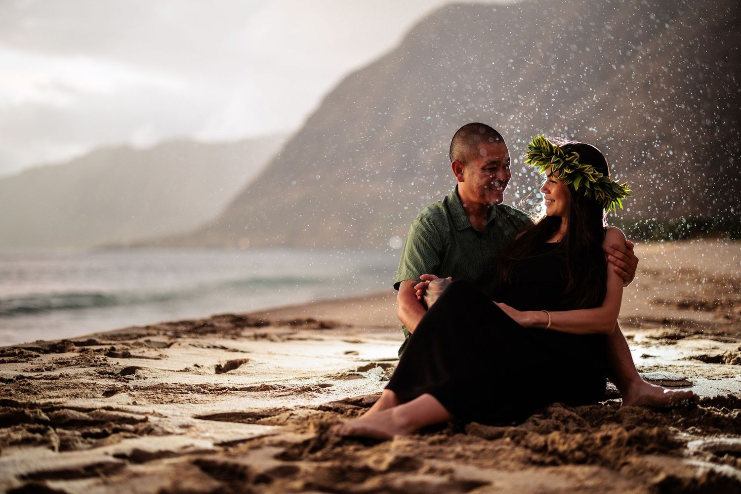 Couple sitting together on a sandy Hawaii beach with misty mountains behind them.