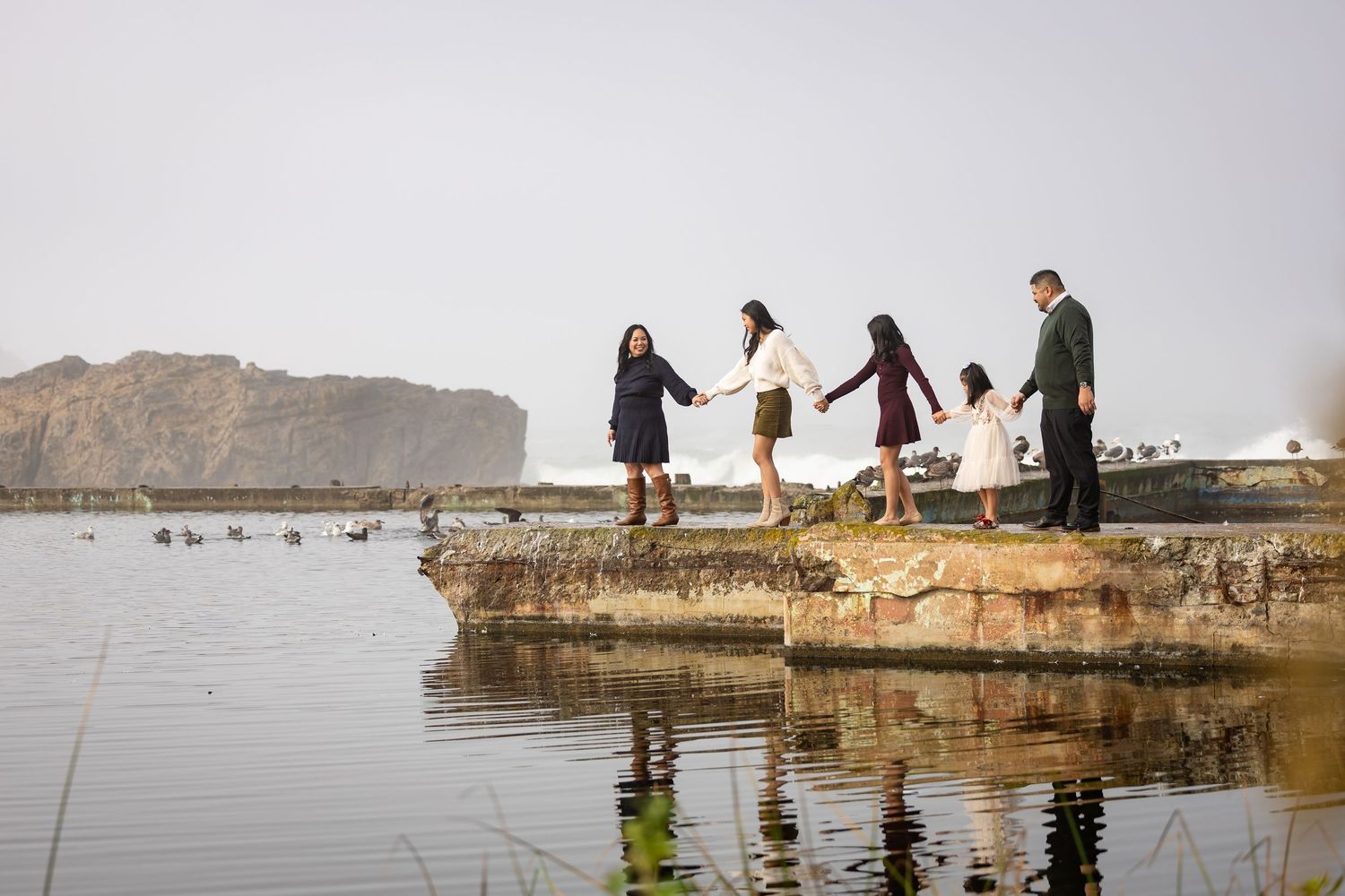 A family holds hands while walking along a rocky waterfront with mountains and calm water in background.