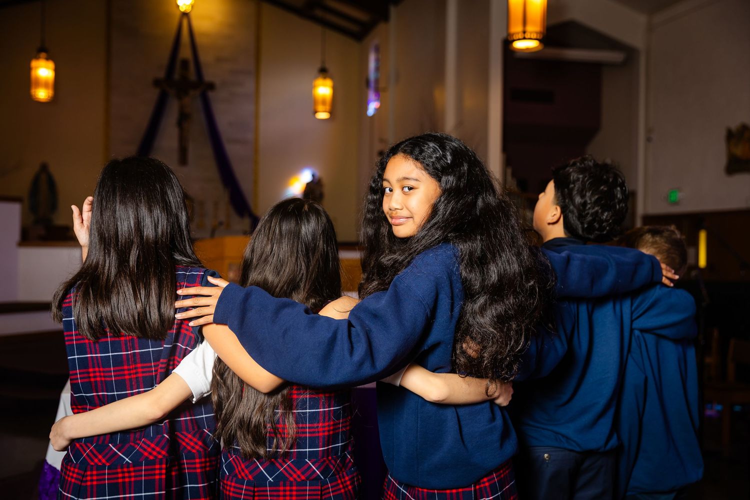 Four girls in school uniforms embracing each other in a church setting.