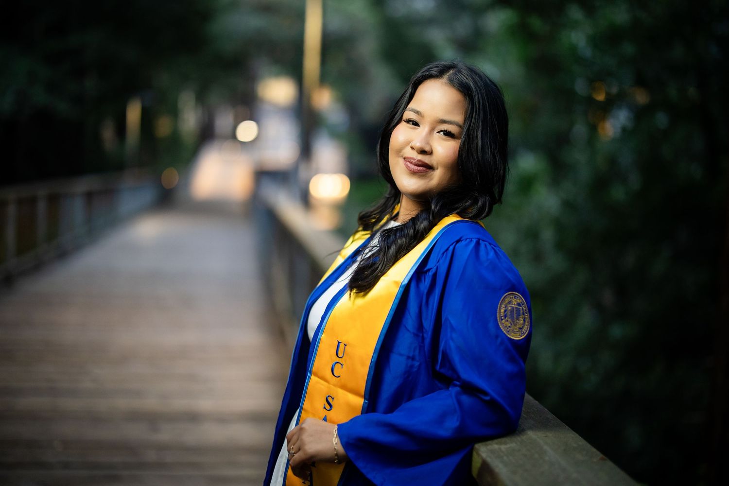 A smiling graduate in blue cap and gown poses on a wooden bridge at dusk for senior photos.