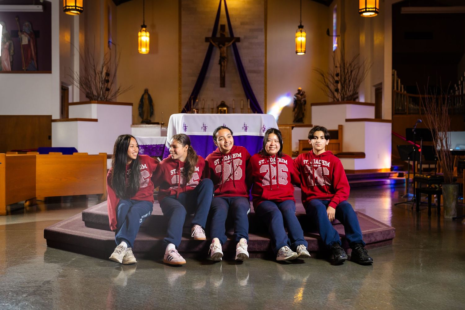 Five students in matching red hoodies sitting together in front of a church altar.