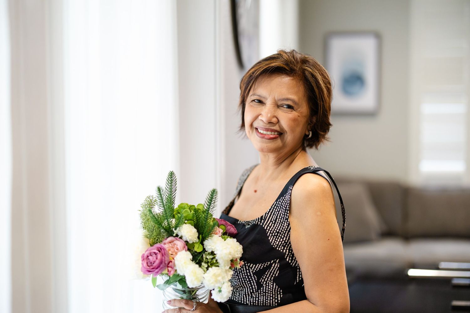 Smiling woman holding a bouquet of pink and white flowers indoors near a bright window.