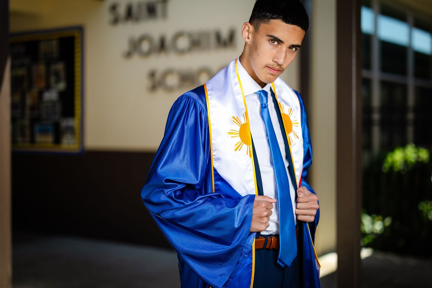 Male graduate in blue cap and gown outside Saint Joachim School with Filipino sash.