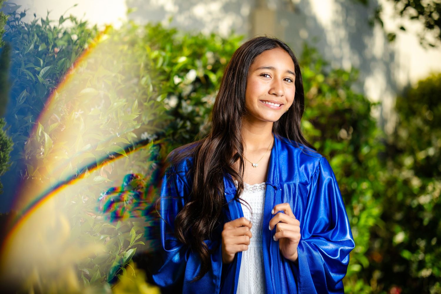 Smiling girl in blue graduation gown standing outdoors with colorful bokeh effect.