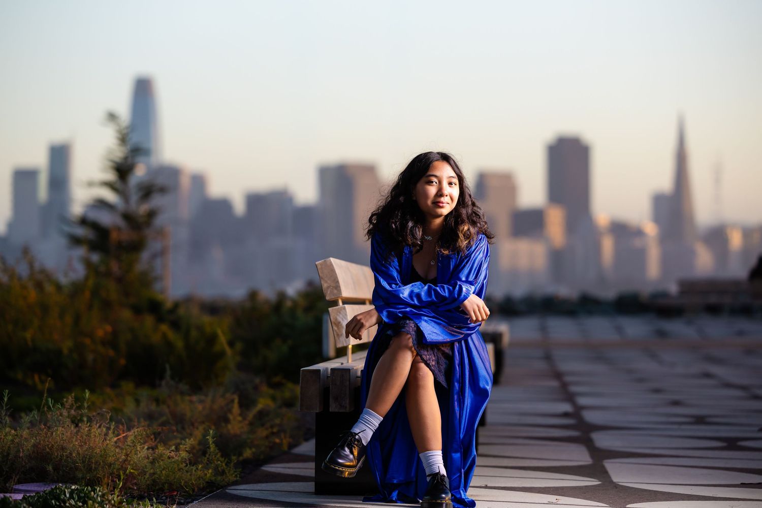 Young woman in blue dress sitting on bench with San Francisco skyline at sunset.