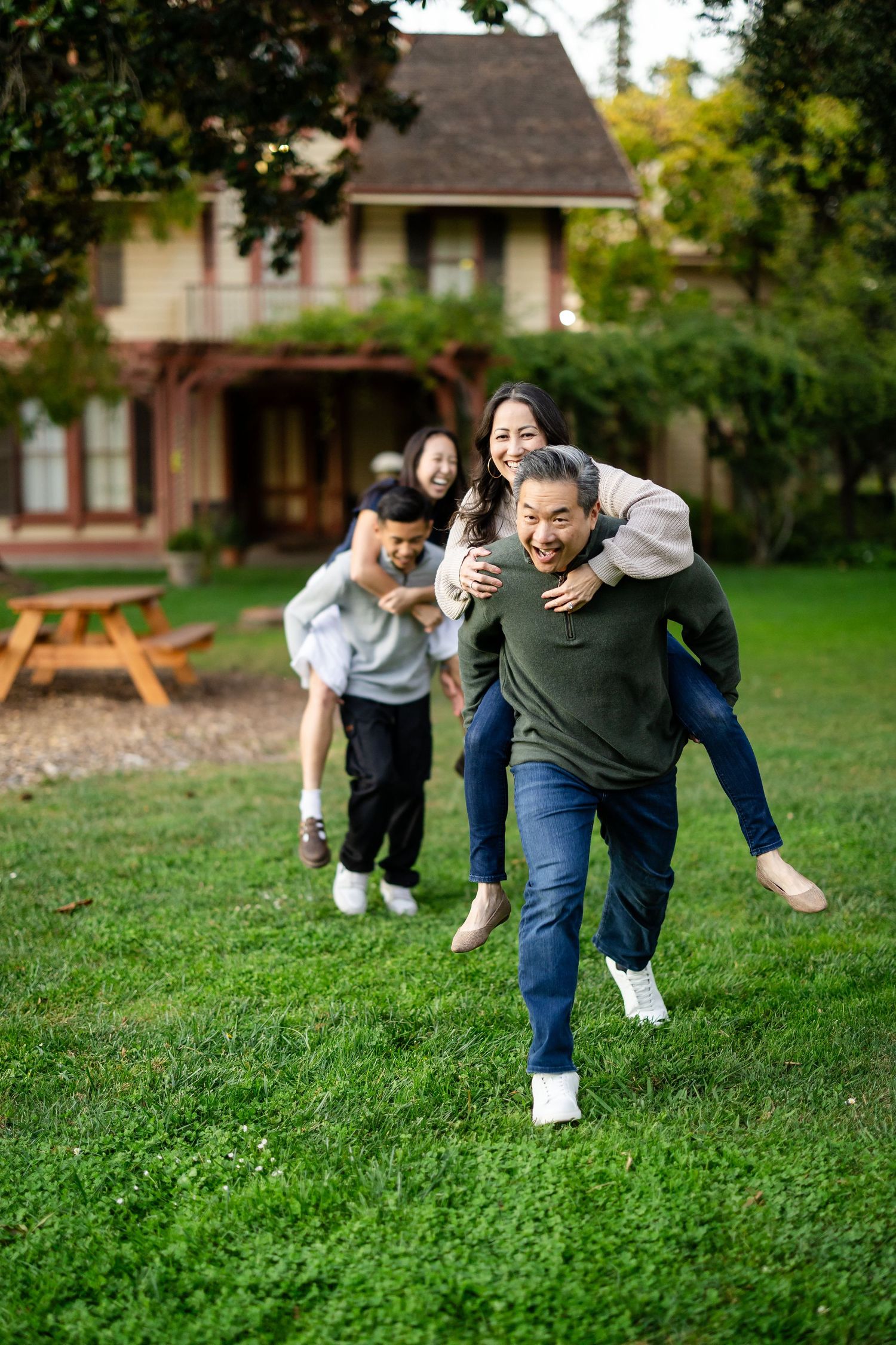 A joyful Asian family plays piggyback in a lush green yard in front of a charming Victorian home.
