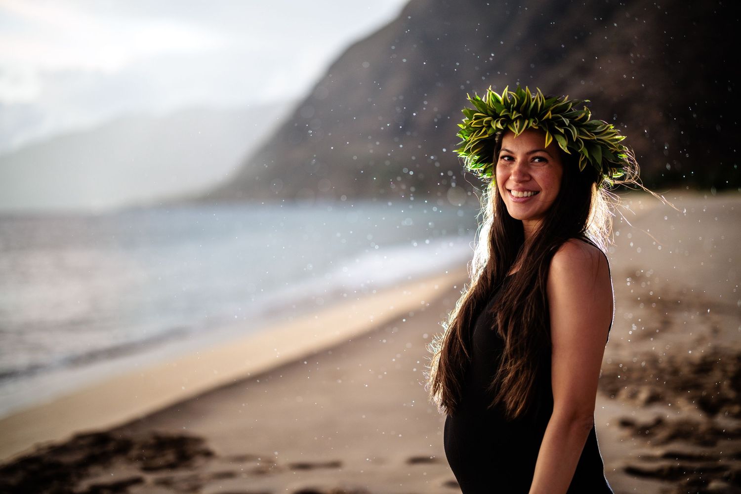 Smiling woman wearing a green leaf crown on a Hawaii beach with mountains in the background.
