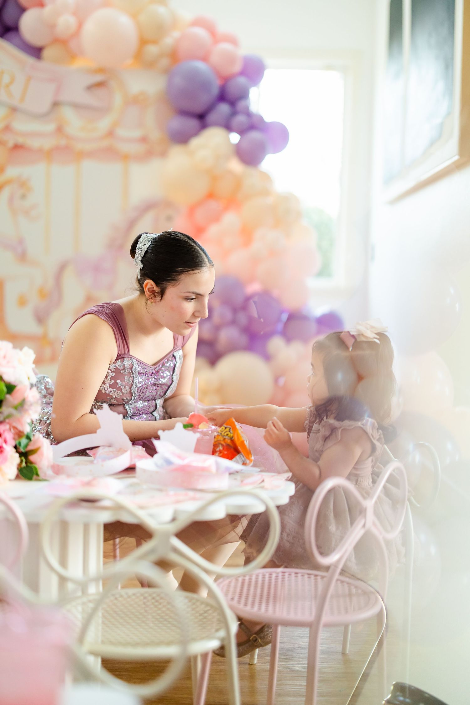 Woman in purple dress interacting with a young girl at a pink party table with balloon decorations.