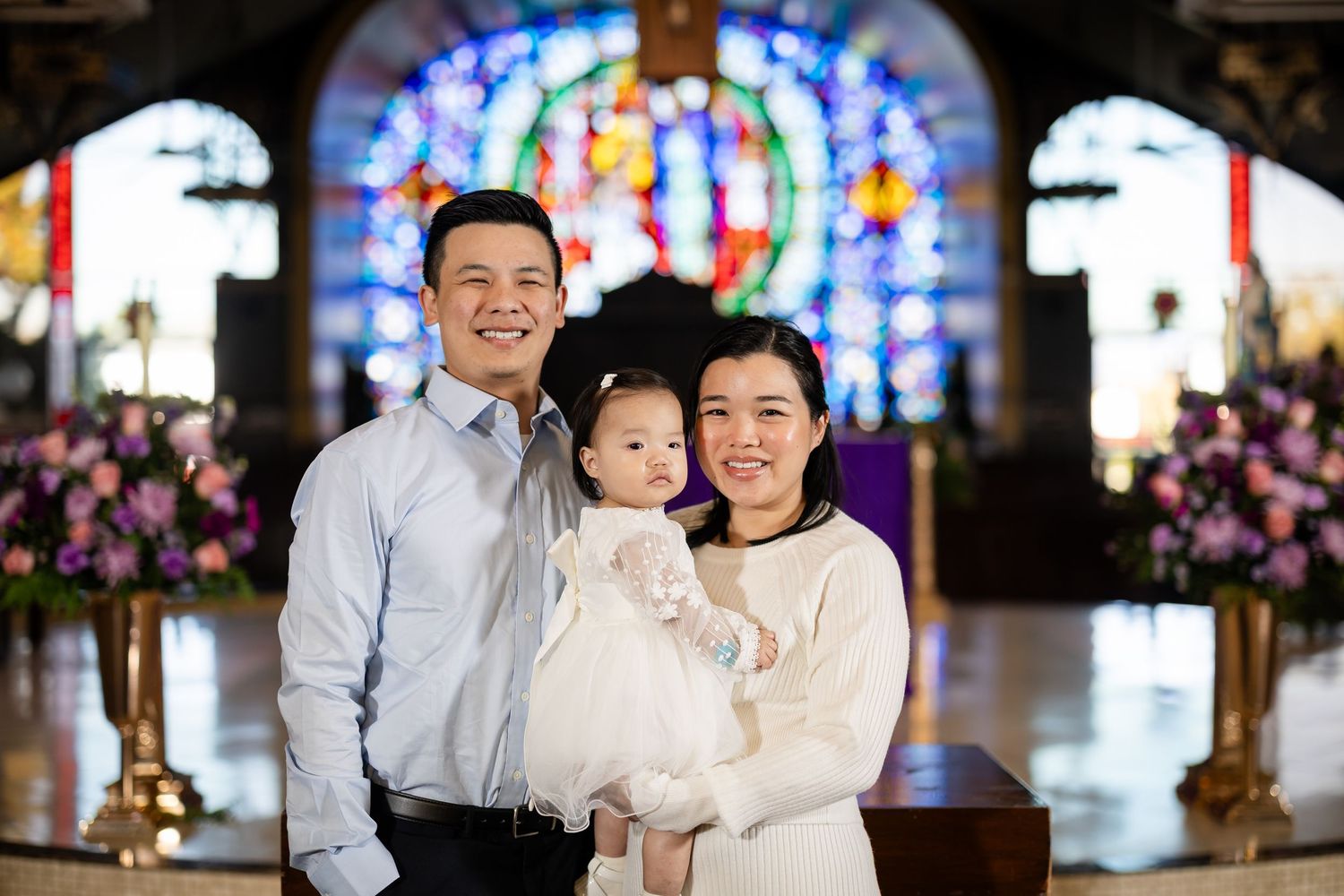 A happy family poses with their baby in white dress inside a church with colorful stained glass windows.