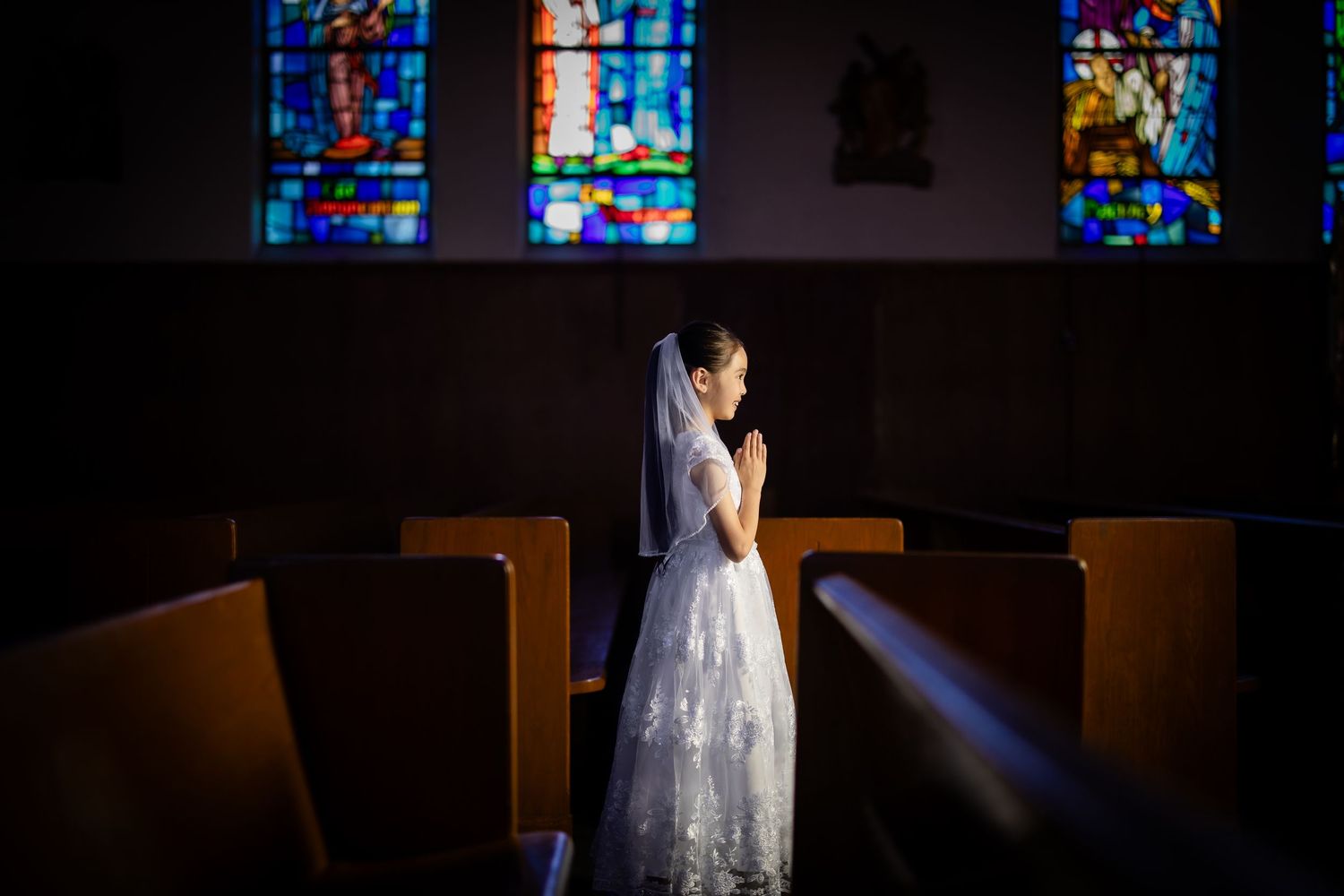 A young girl in a white First Communion dress and veil prays in a sunlit church pew.