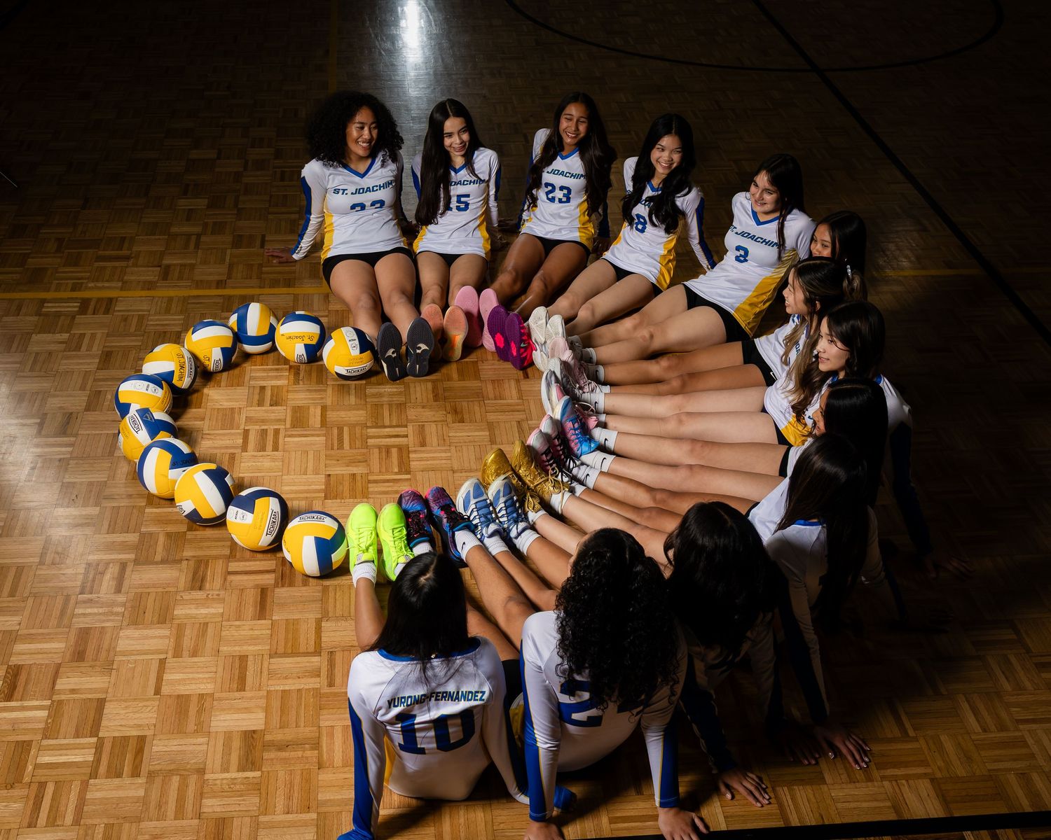 A girls volleyball team forms a heart shape with volleyballs on a hardwood gym floor, aerial view.
