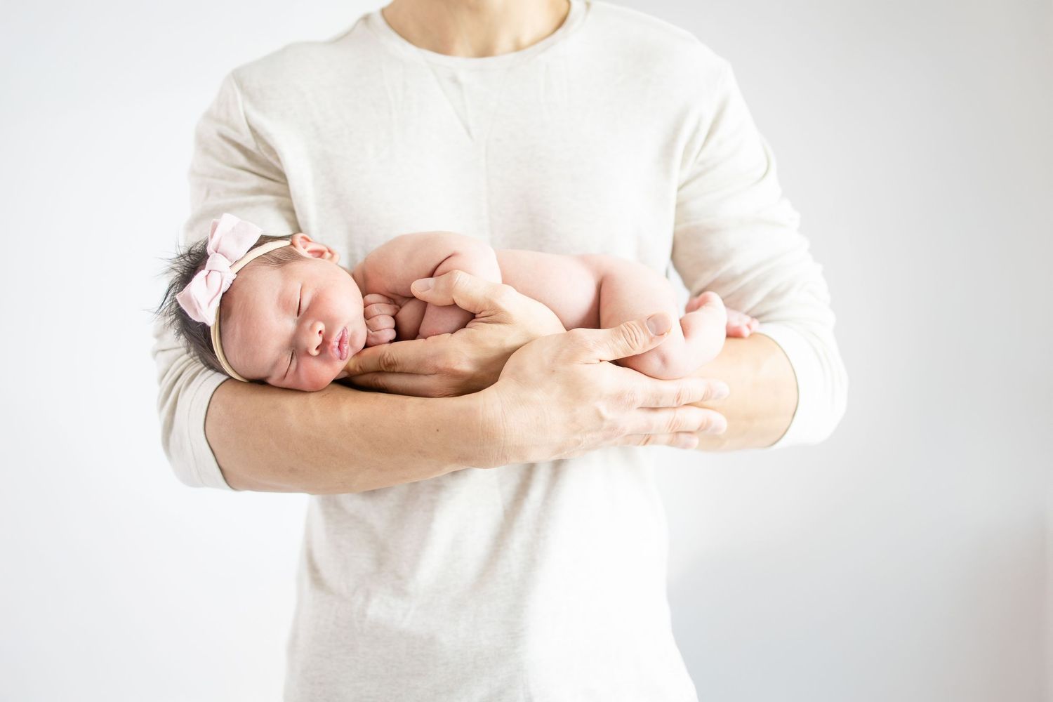 A newborn baby in a pink bow sleeps peacefully in a parent's gentle hands against white background.