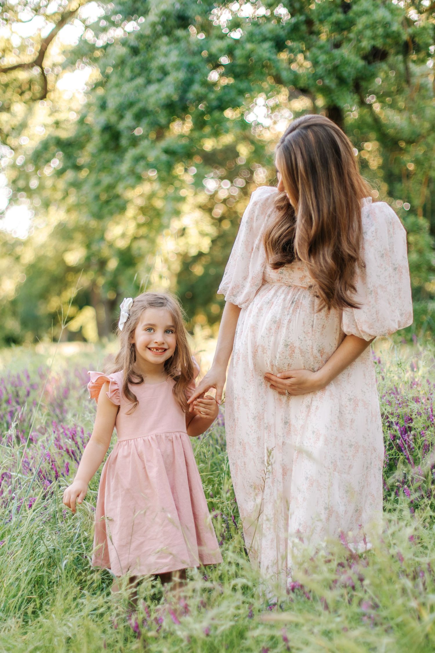 Young girl in pink dress holding hands with her pregnant mother in a sunlit flower field.
