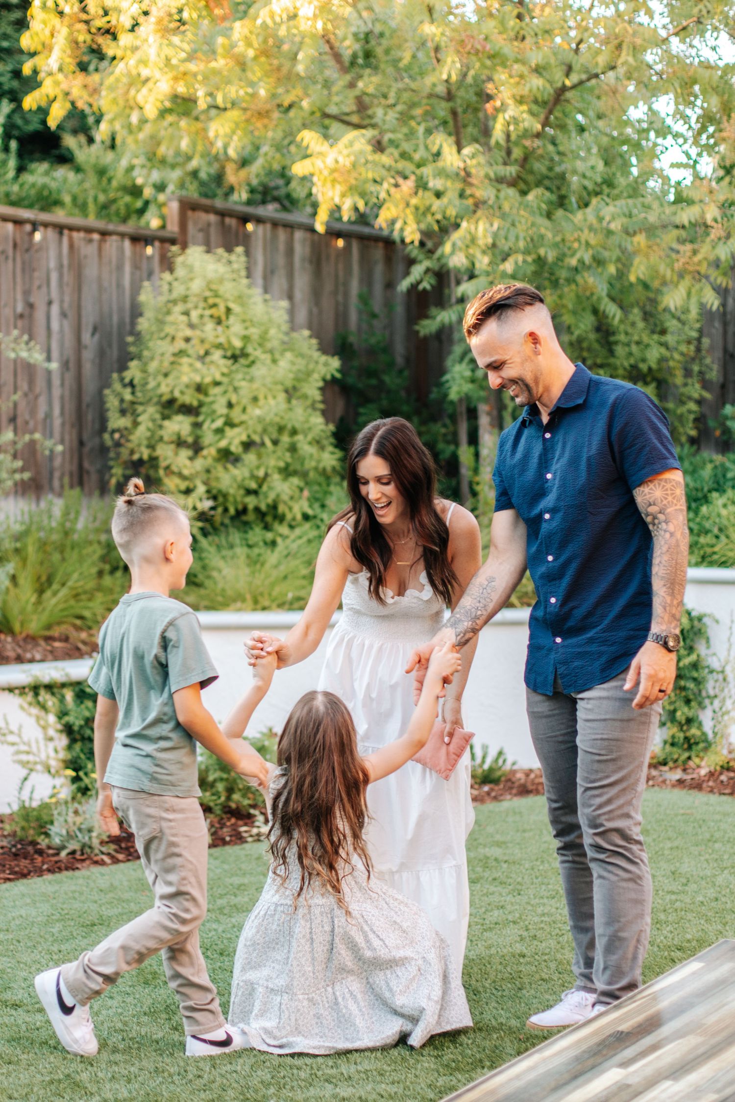 A tattooed dad and mom laugh with two children while playing together on a green backyard lawn.