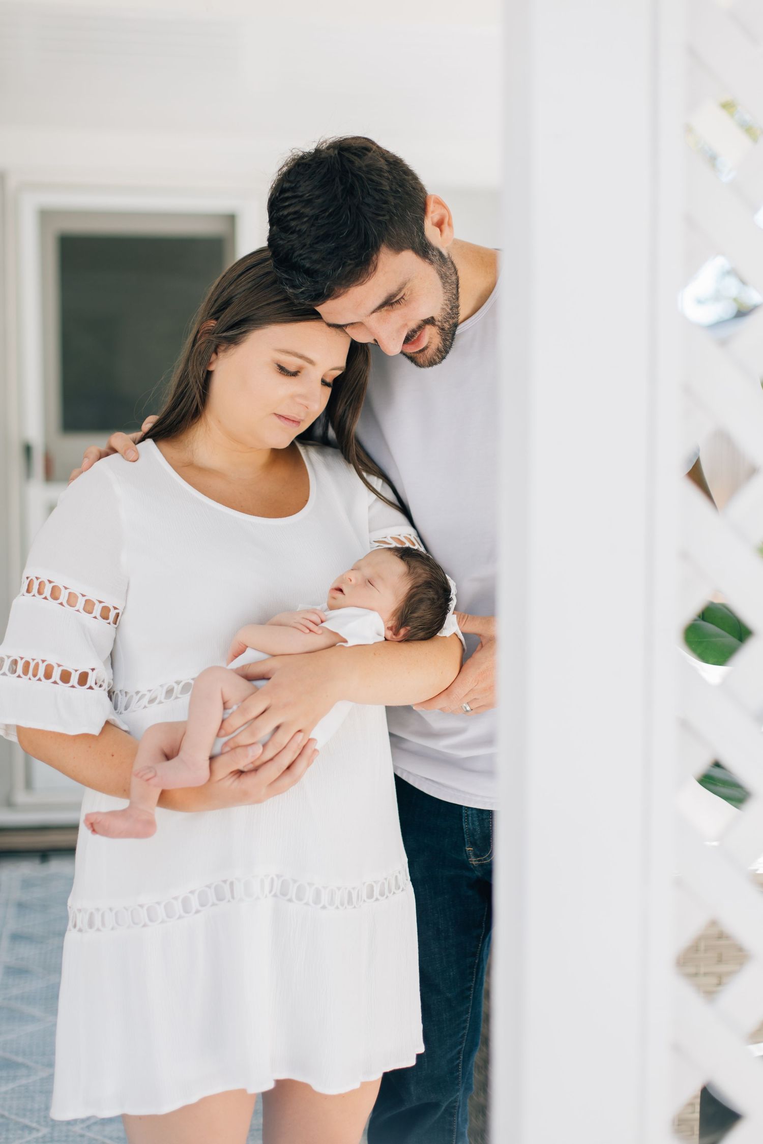 Young couple in white clothing tenderly hold their sleeping newborn baby on a porch.