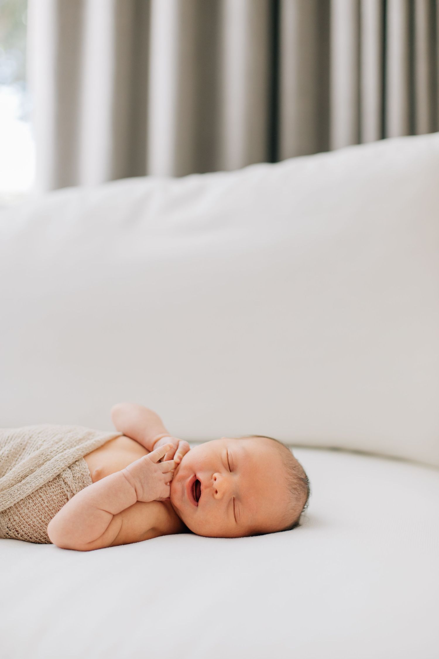 Newborn baby in beige wrap lies on white bed with mouth open in a yawn.