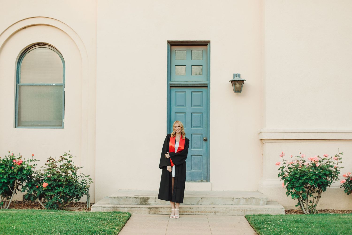 Graduate in black cap and gown holding diploma outside a building with a teal door and rose bushes.