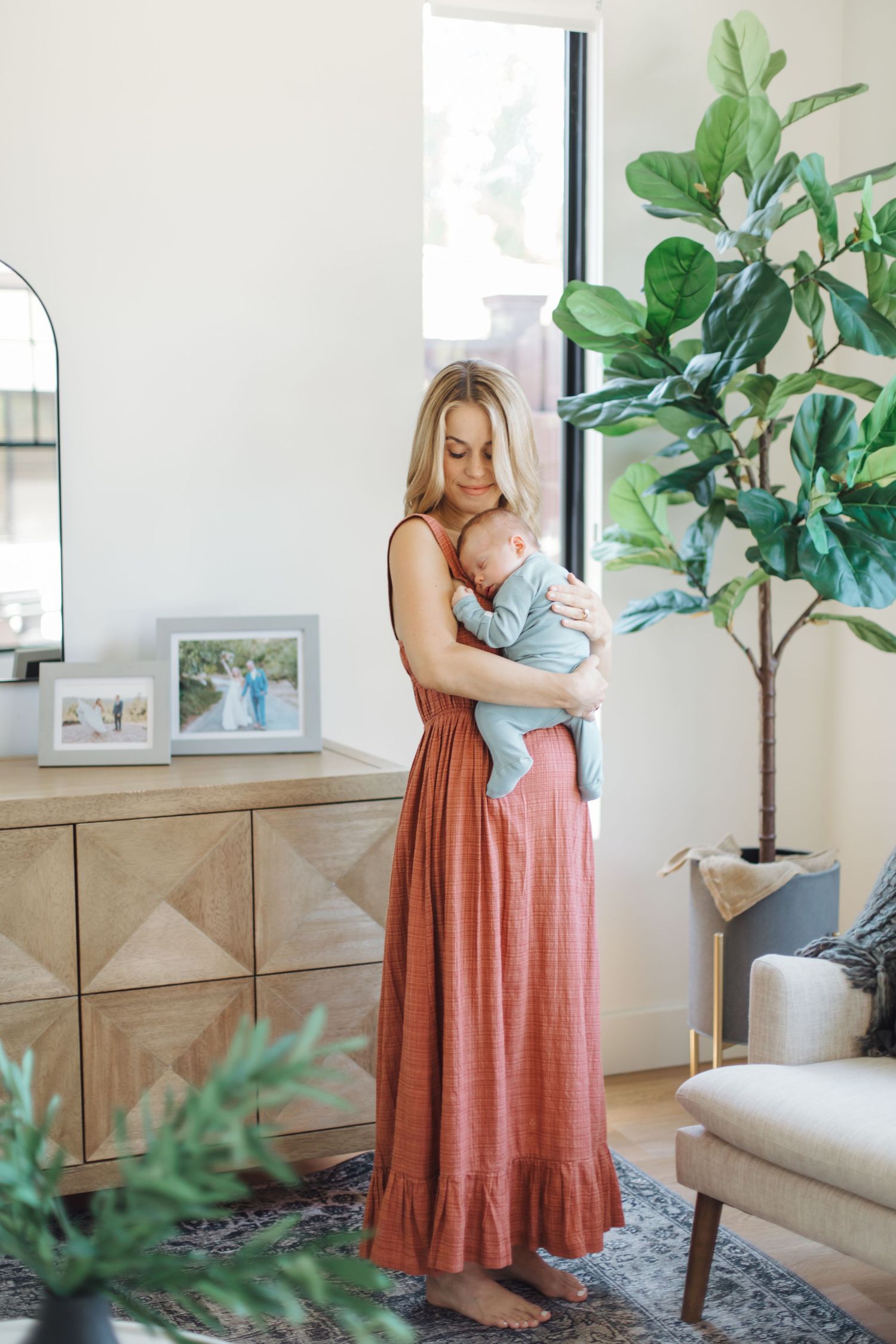 Full-length portrait of mother in terracotta maxi dress holding newborn in bright living room.