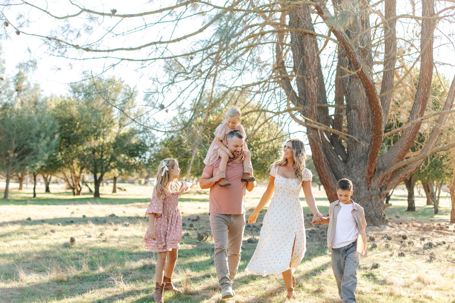 Family of five walking through a sunny park, parents holding hands with children, one child on dad's shoulders.