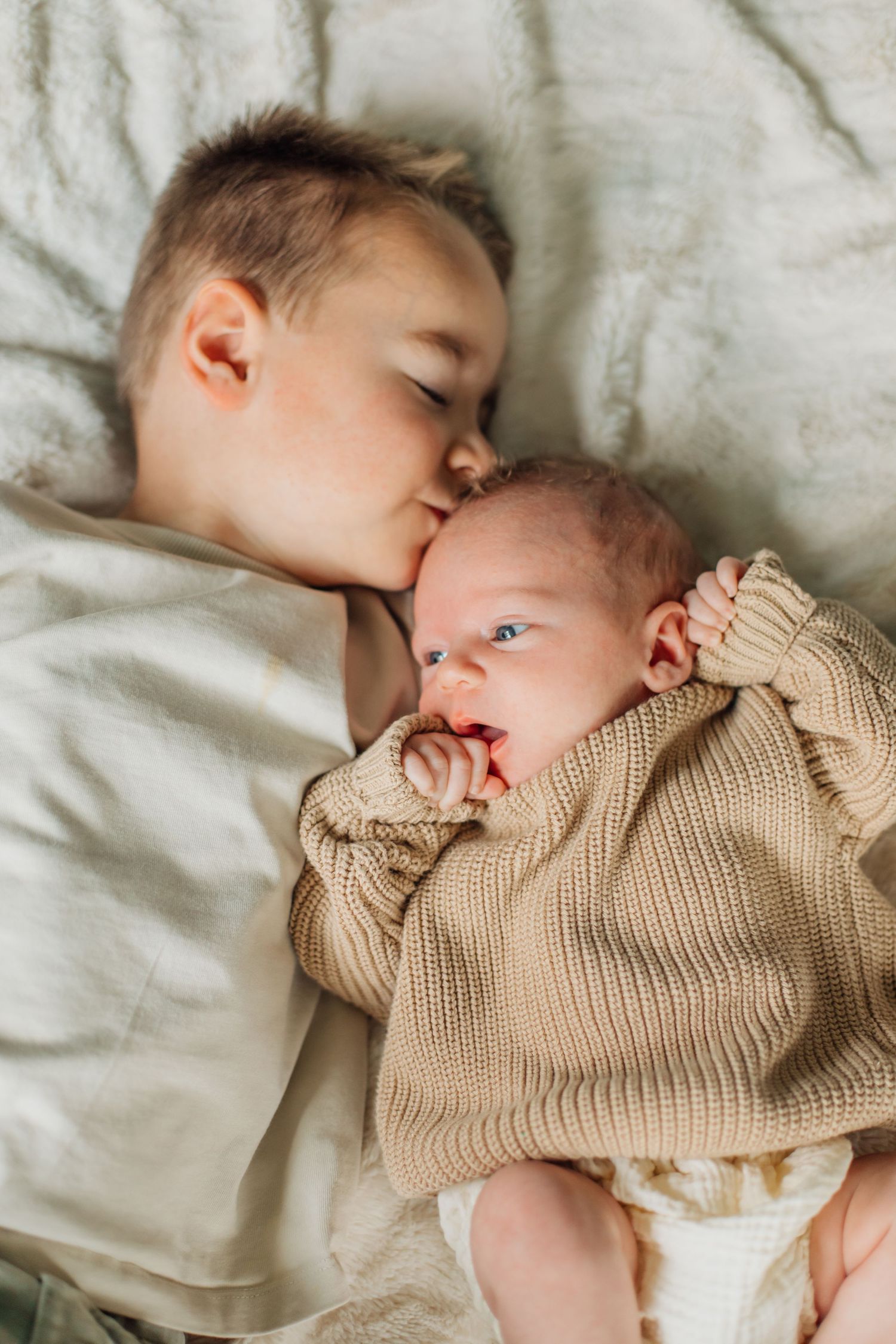 Young boy kissing newborn sibling lying on white bedding wearing beige knit sweater.