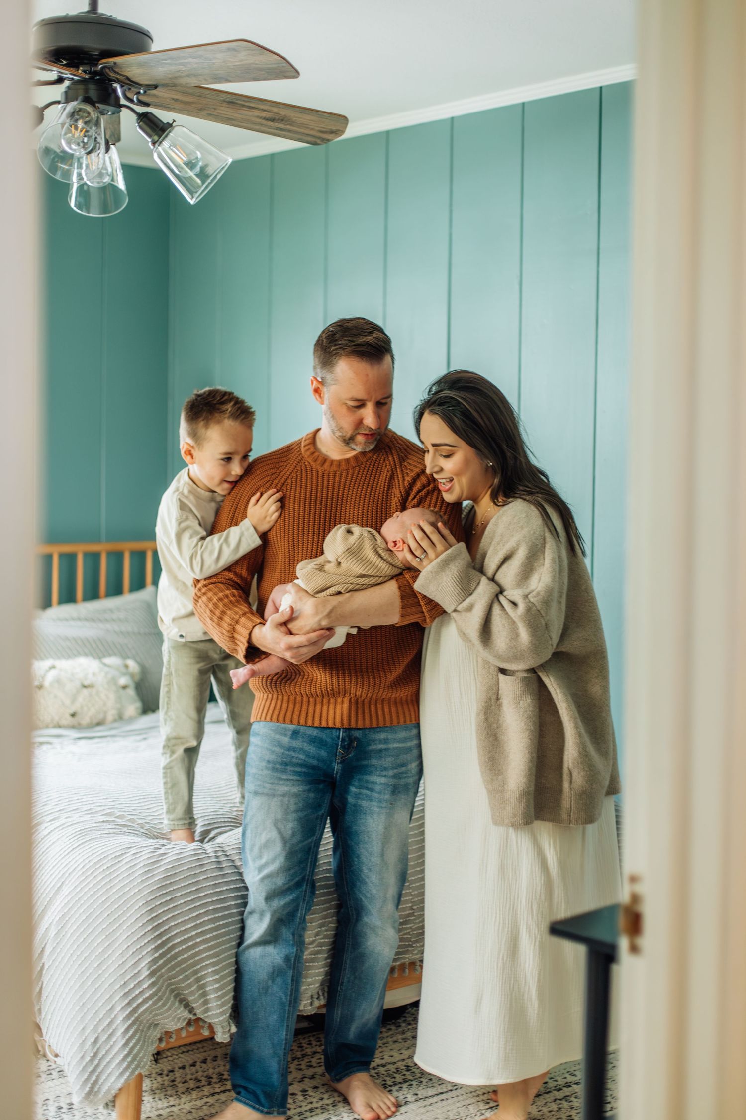 A happy family of four gathers in a teal bedroom, parents and son admiring their newborn baby.
