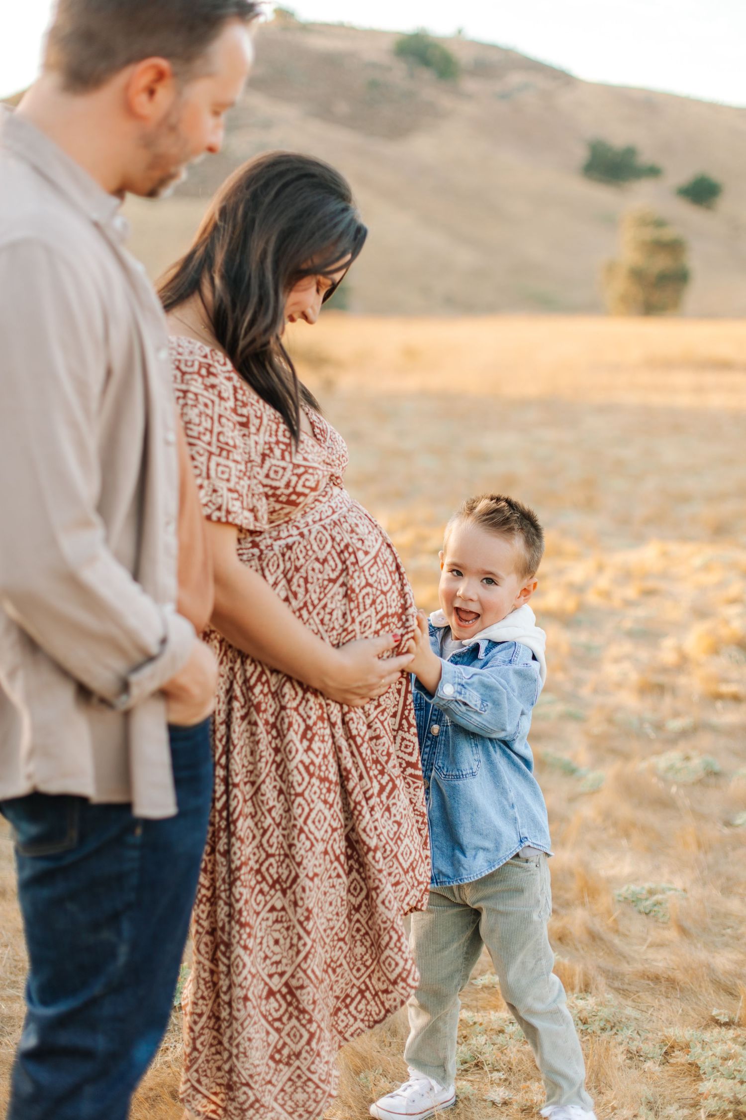 Happy toddler boy in denim shirt smiling at camera while parents cradle pregnant belly outdoors.