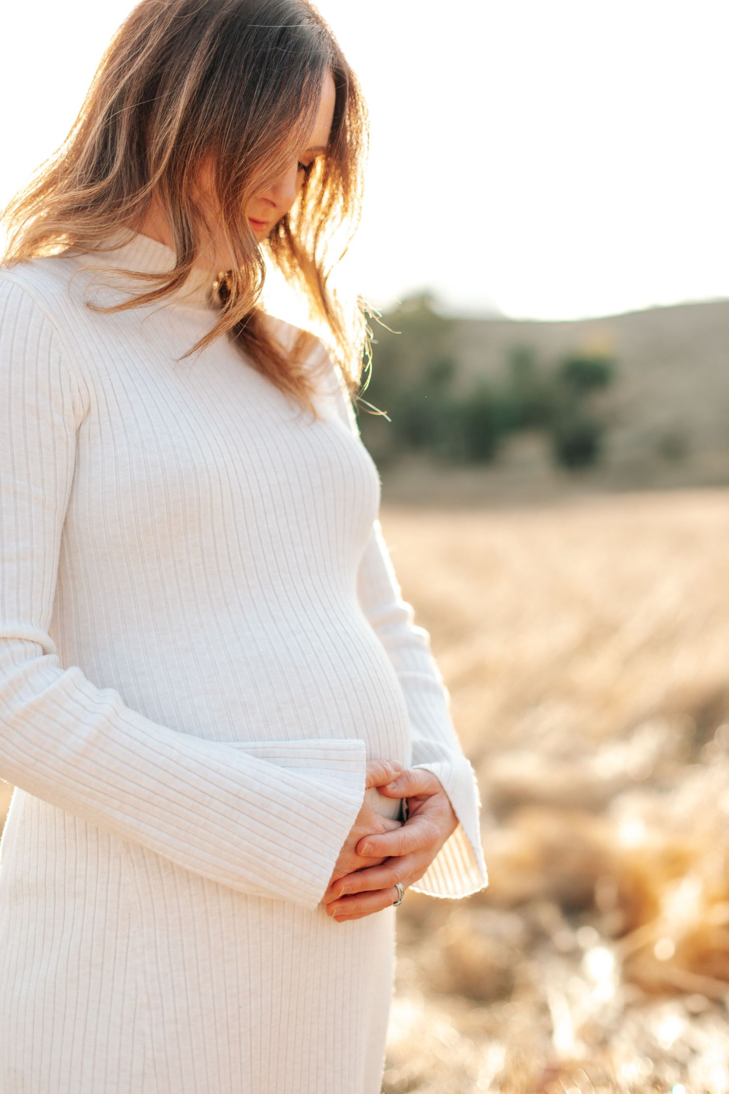 Pregnant woman in white ribbed sweater dress cradling her baby bump in a golden field at sunset.