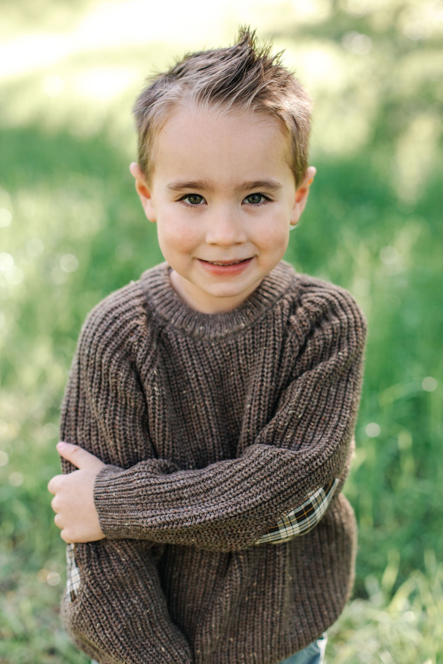 Adorable young boy in a brown knit sweater smiling with arms crossed in a green outdoor setting.