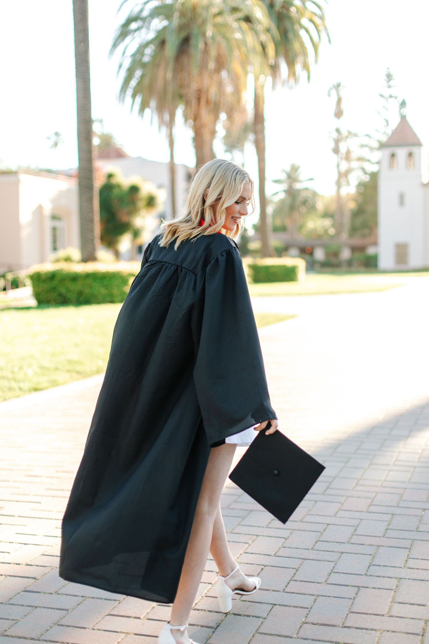 Blonde woman in black graduation gown holding cap while walking on sunny brick path near palm trees.