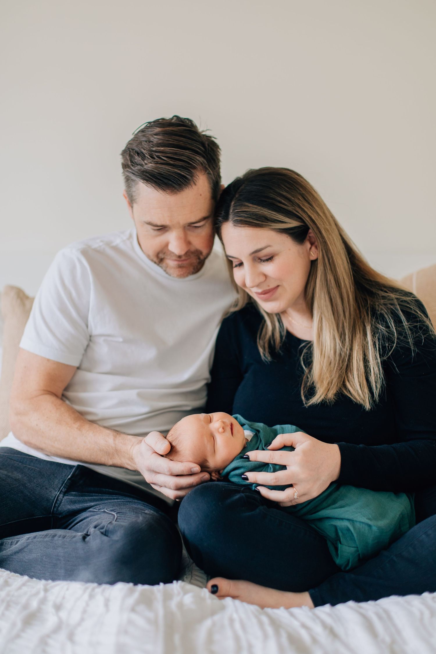New parents sitting on bed smiling lovingly at their newborn baby wrapped in teal blanket.