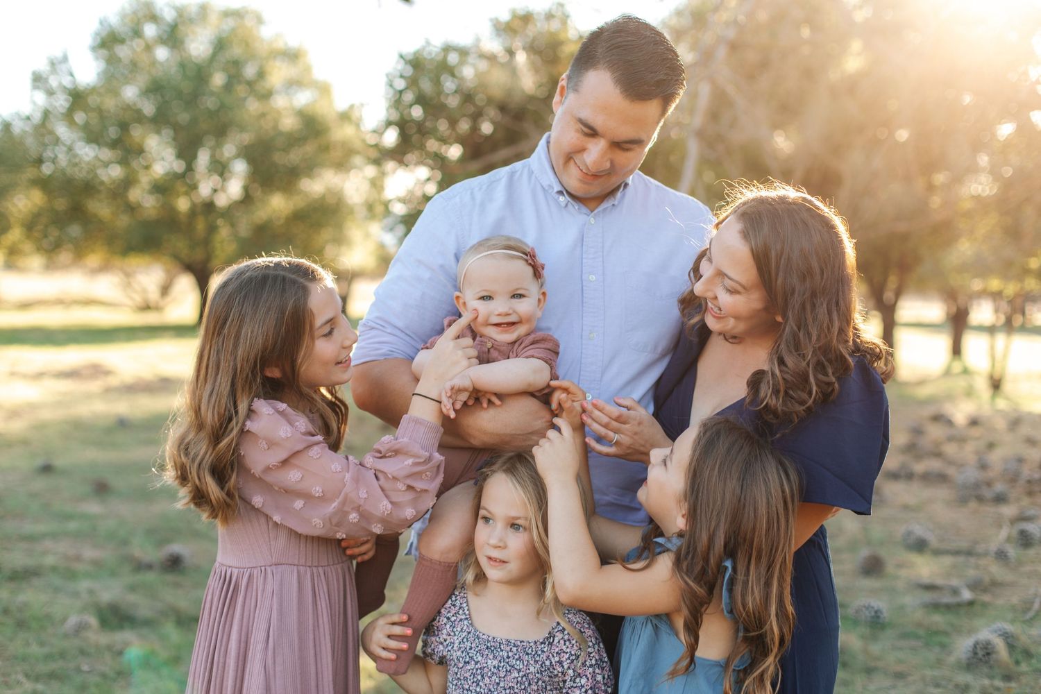 Happy family of six with baby and four daughters gathered together in a sunlit park.