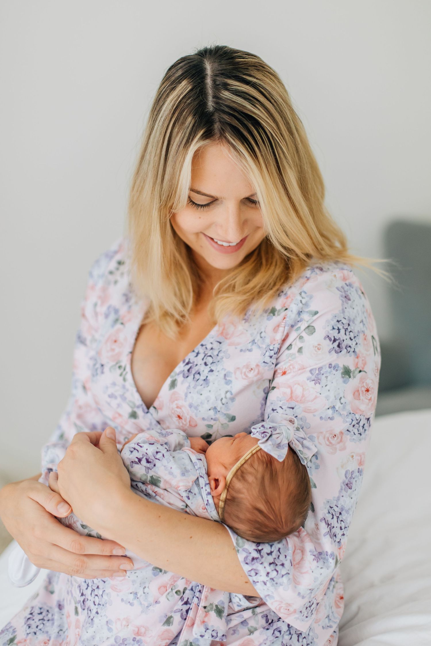 Smiling blonde mother in floral dress tenderly holds her newborn baby while sitting on a bed.