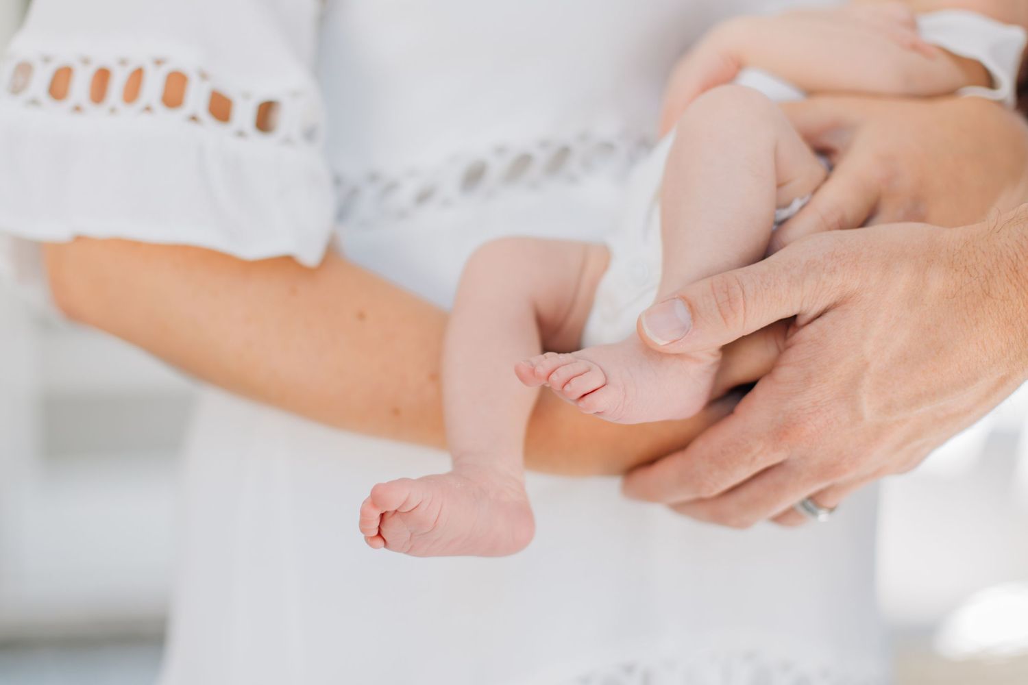 Close-up of parents hands gently holding tiny newborn baby feet in white clothing.