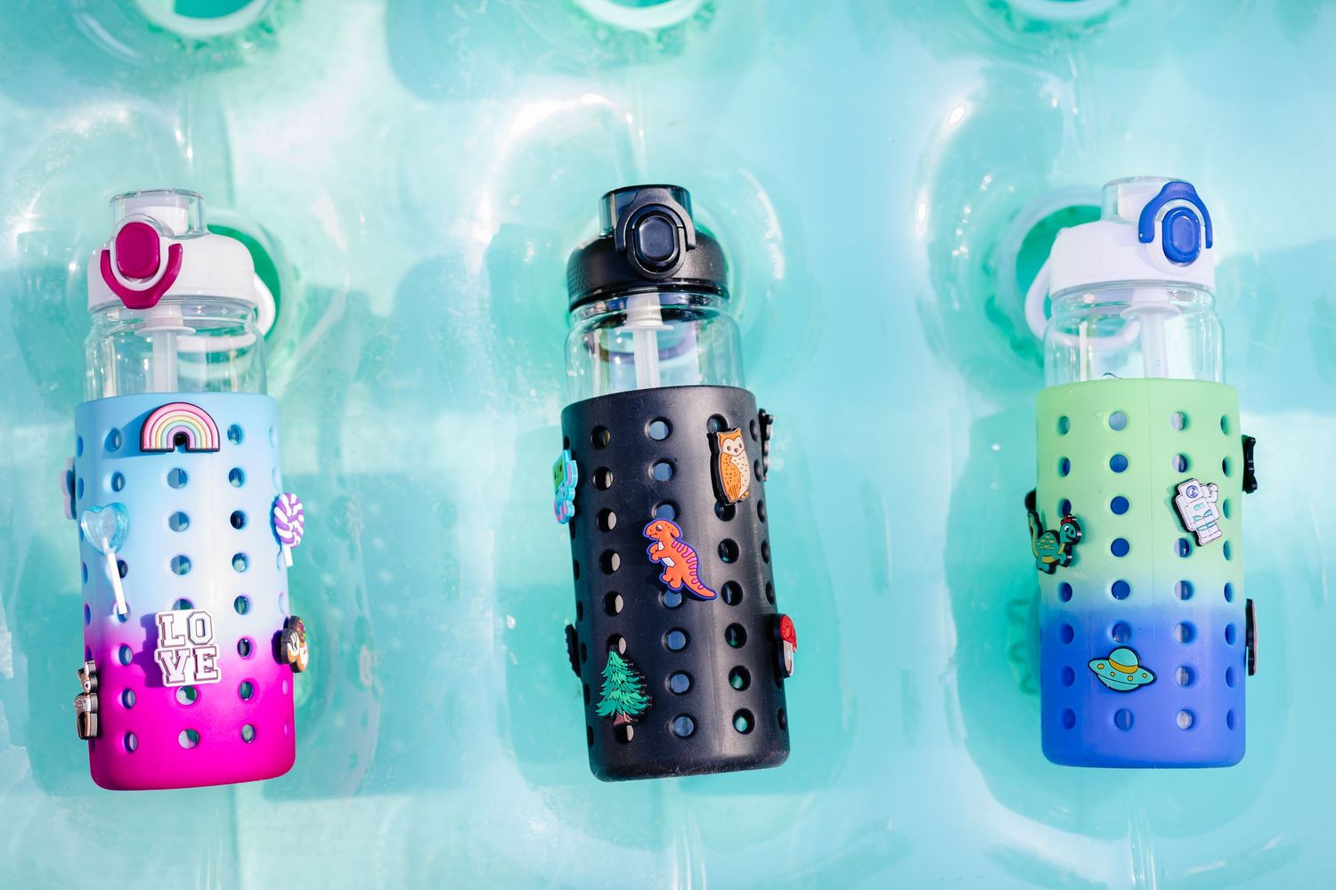 Three colorful charm water bottles in pink, black, and green silicone sleeves on teal background.