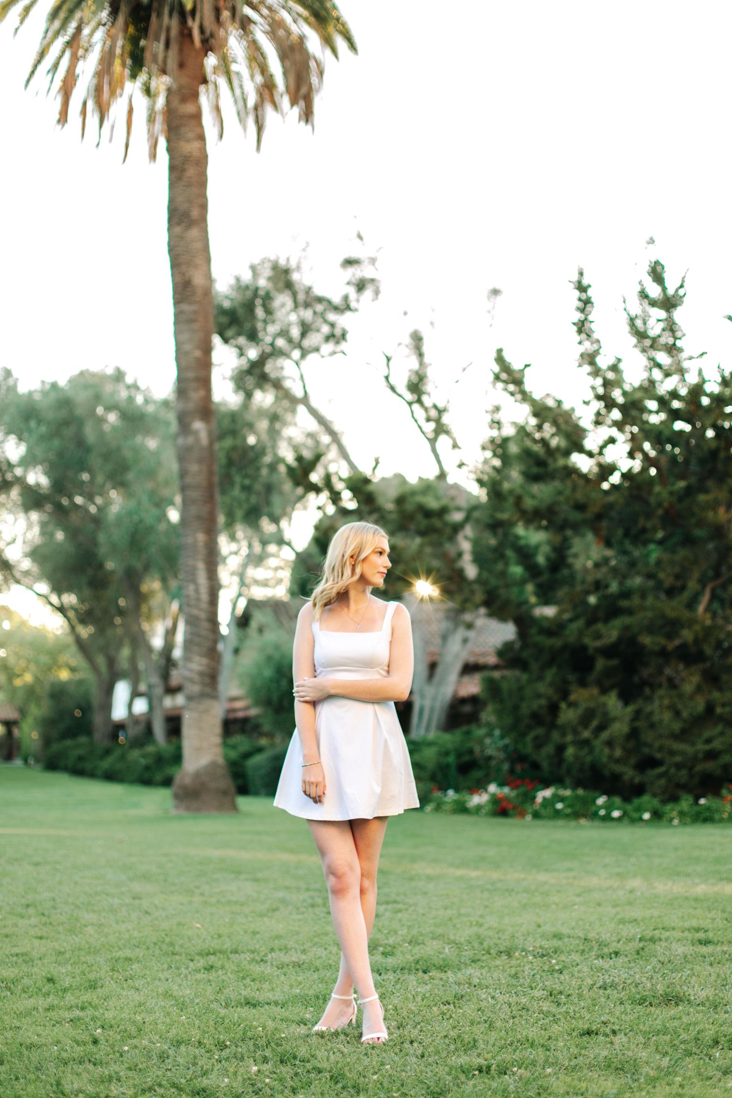 Blonde woman in white mini dress standing on green lawn near a palm tree at golden hour.