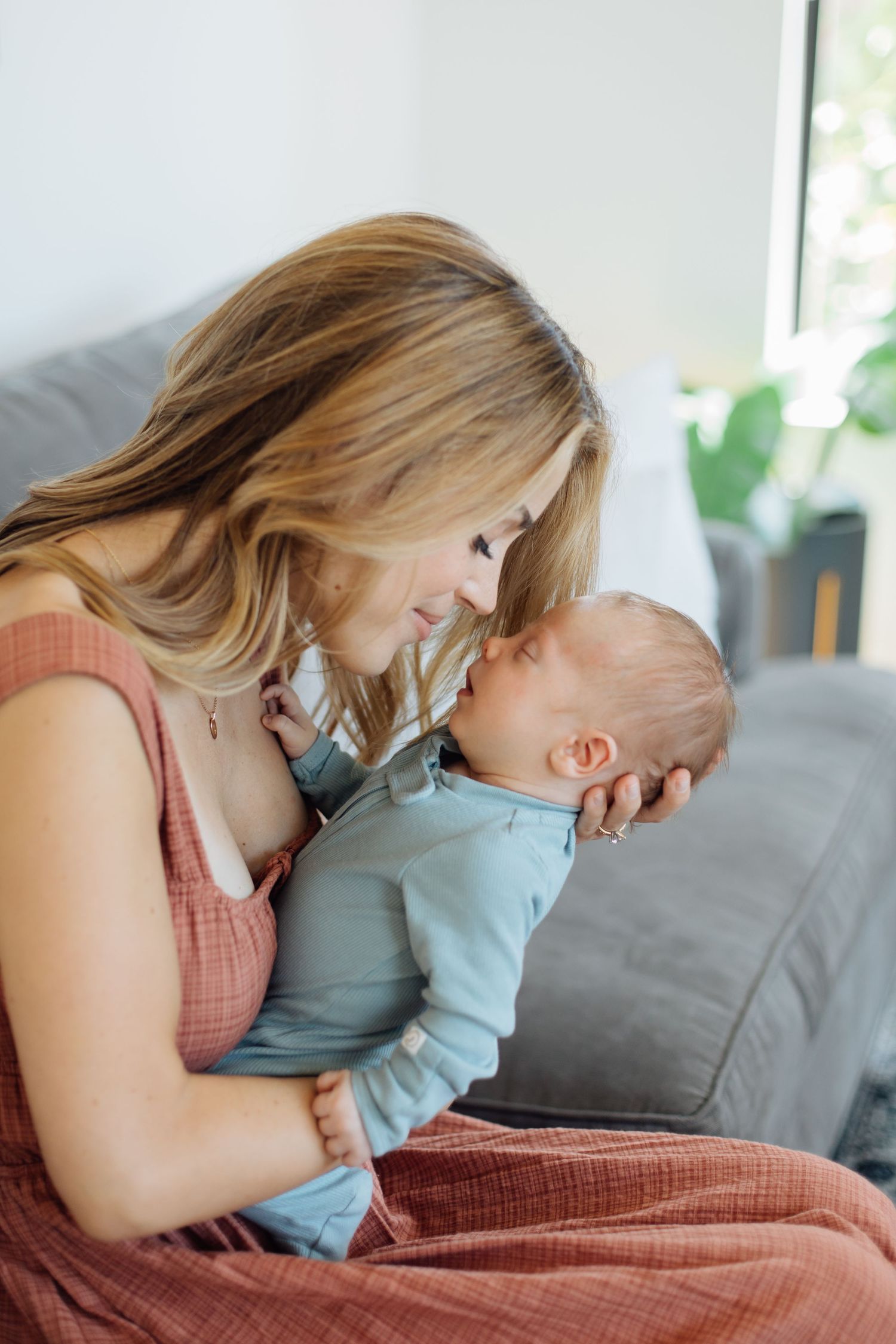 Tender moment of smiling mother sitting on couch holding alert newborn baby in blue onesie.