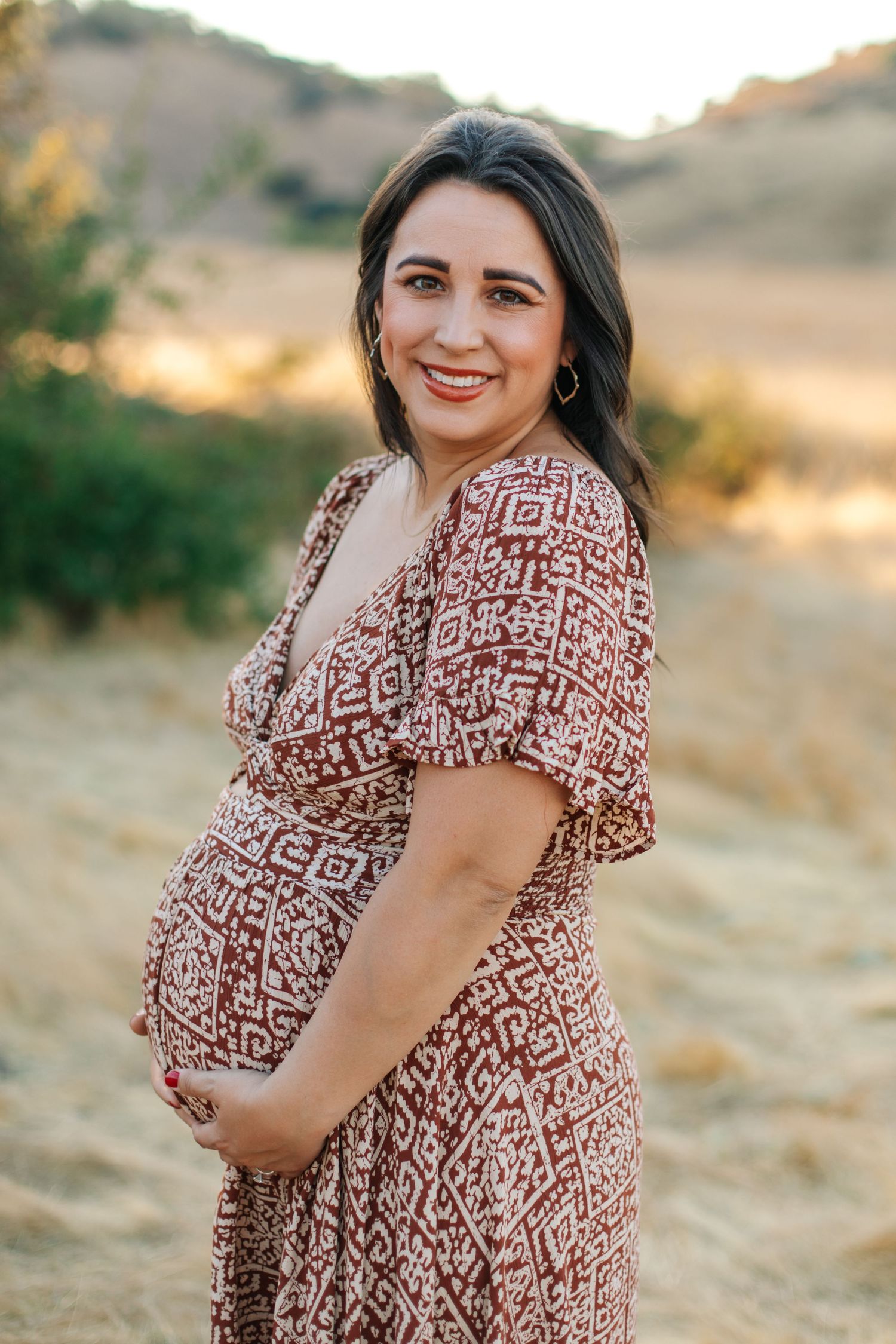 Smiling pregnant woman in red patterned dress holding baby bump in a sun-drenched outdoor field.
